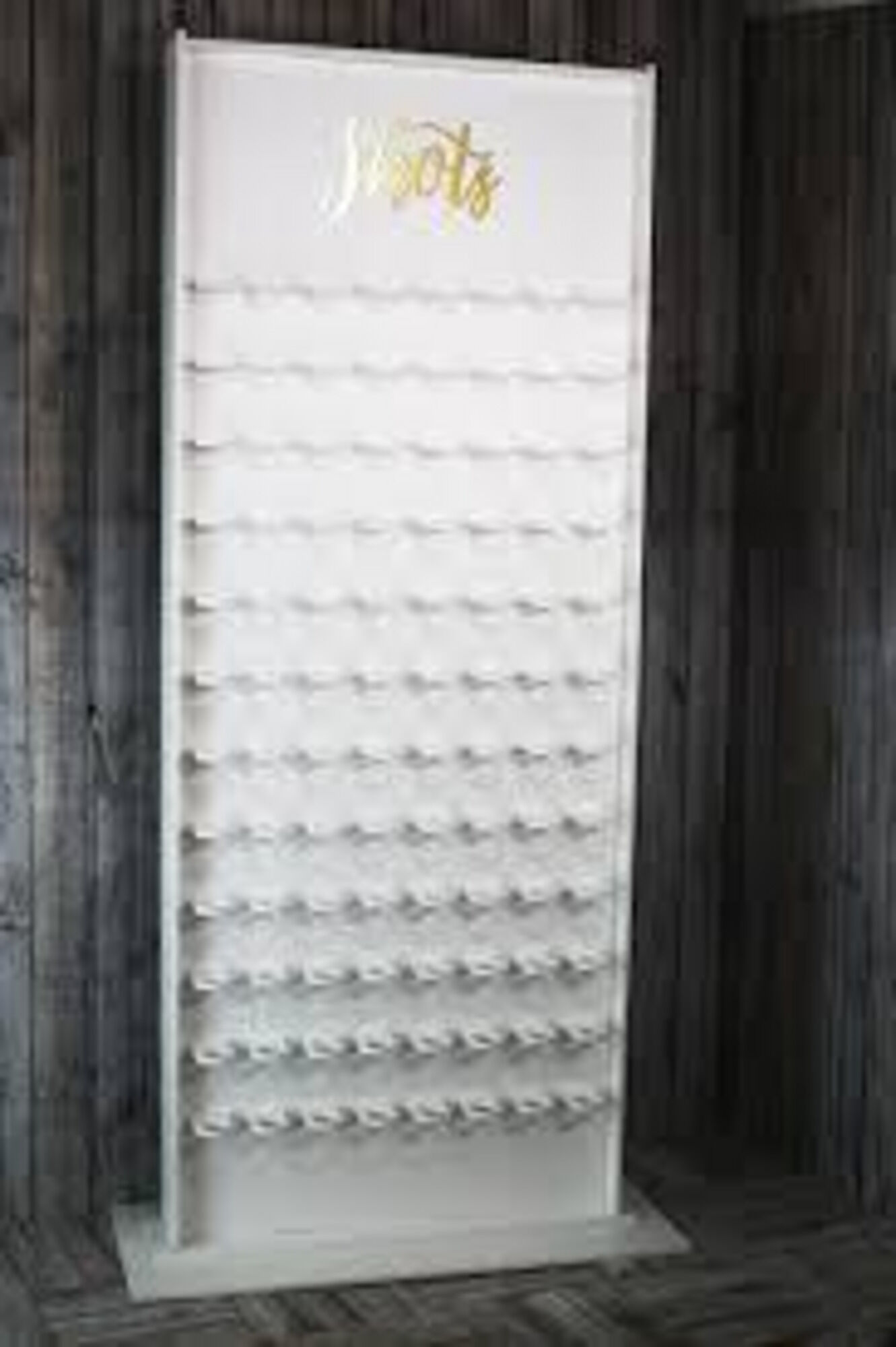 Mur à verre à shooter : panneau rectangulaire blanc posé sur un socle plat, lettrage doré en cursive Shots dans le haut, rangées d’encoches blanches sur toute la hauteur, devant un mur de planches sombres