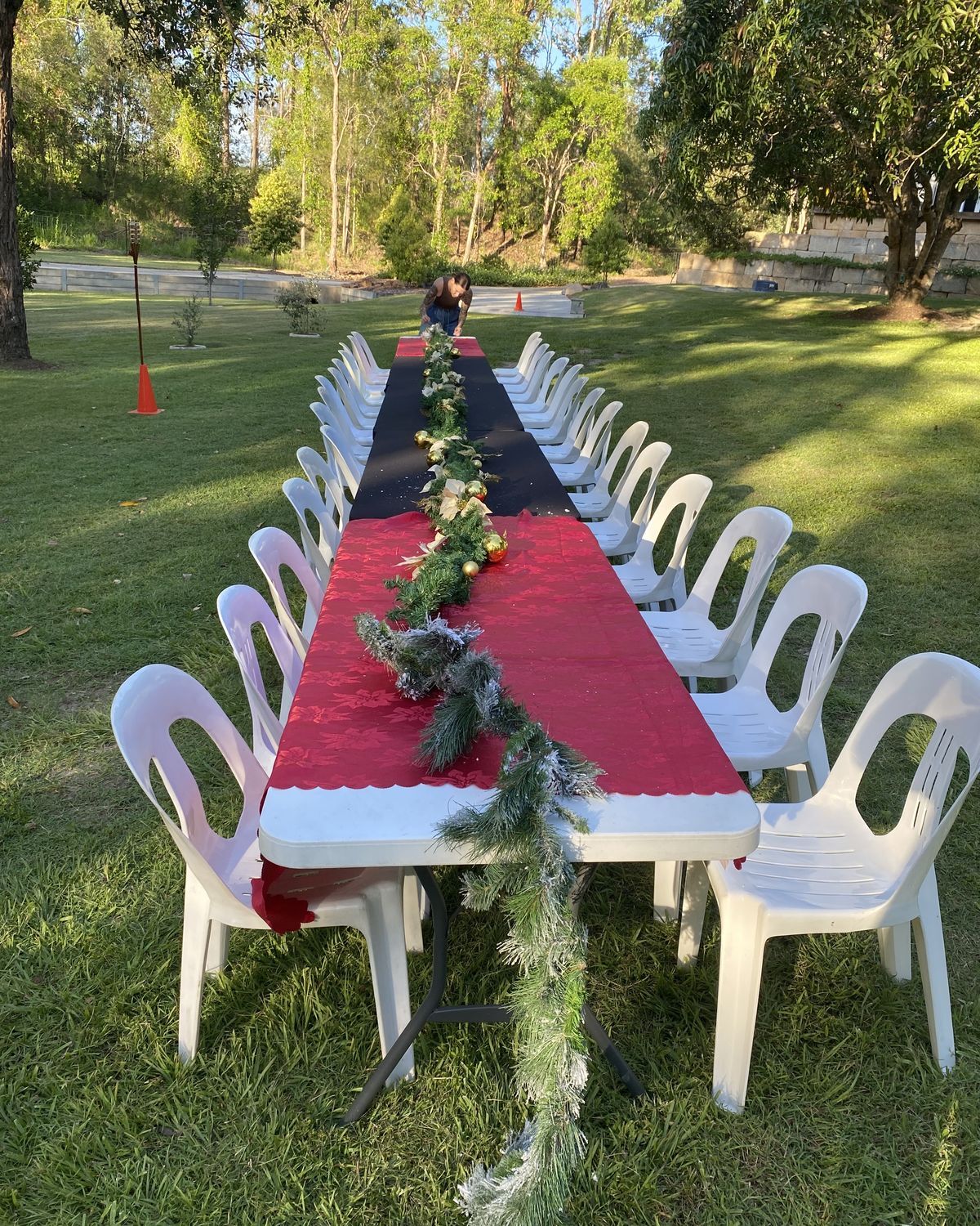 Long outdoor table with a red Christmas runner and garland, set with white chairs on the grass by Adept Hire